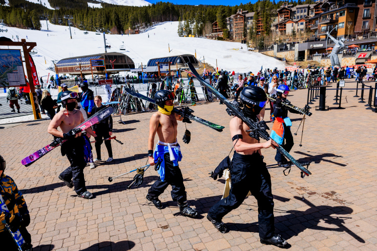 Three shirtless men wearing helmets and goggles carry ski equipment on a stone path at a ski resort.