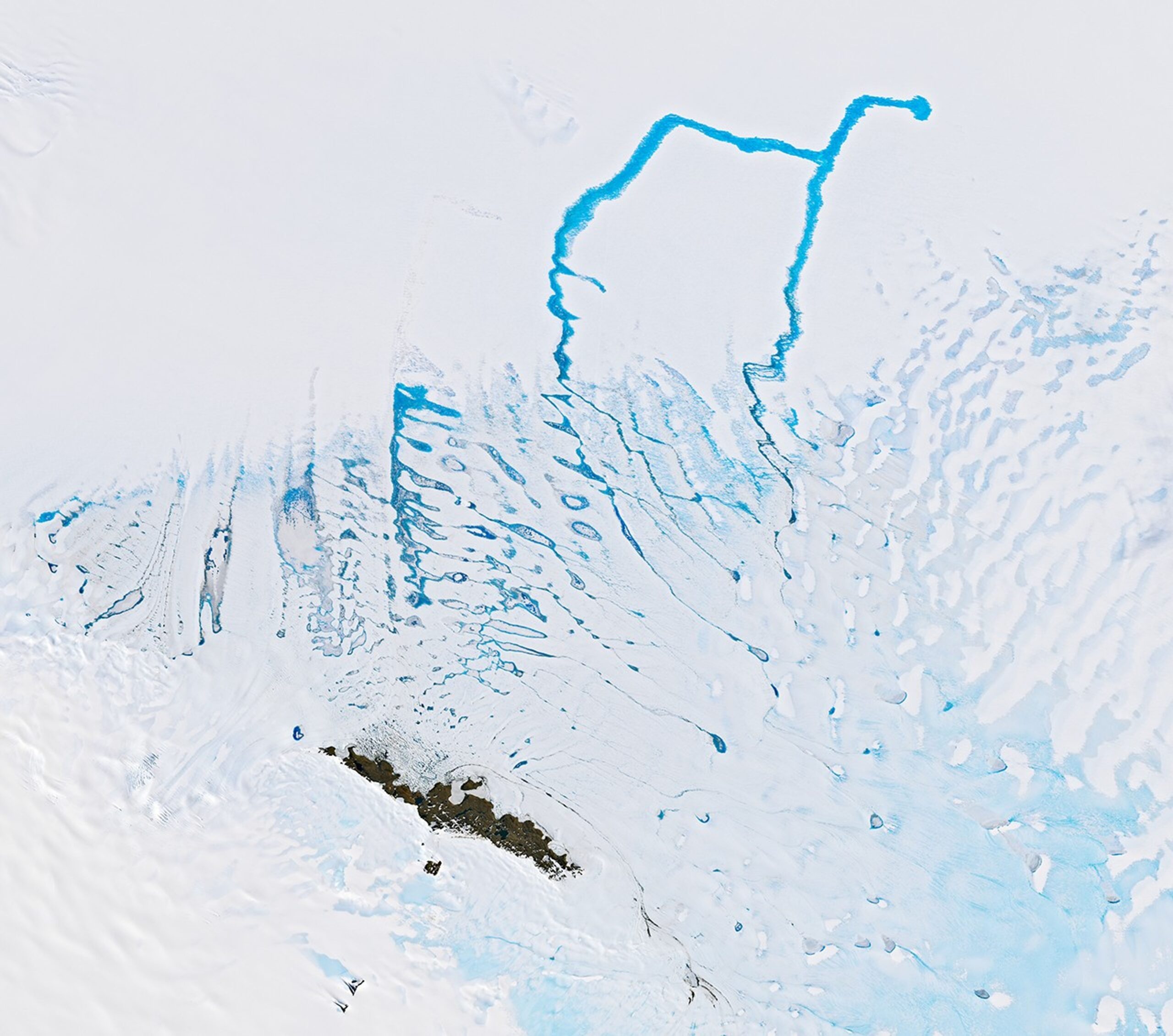 A network of cerulean meltwater channels flowing over the blue-white ice. An "oasis" of the land appears as a brown area of ​​rocks in the lower part of the image.