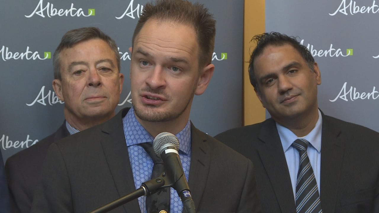 A man in a suit speaks into a microphone while two men in suits stand behind him.