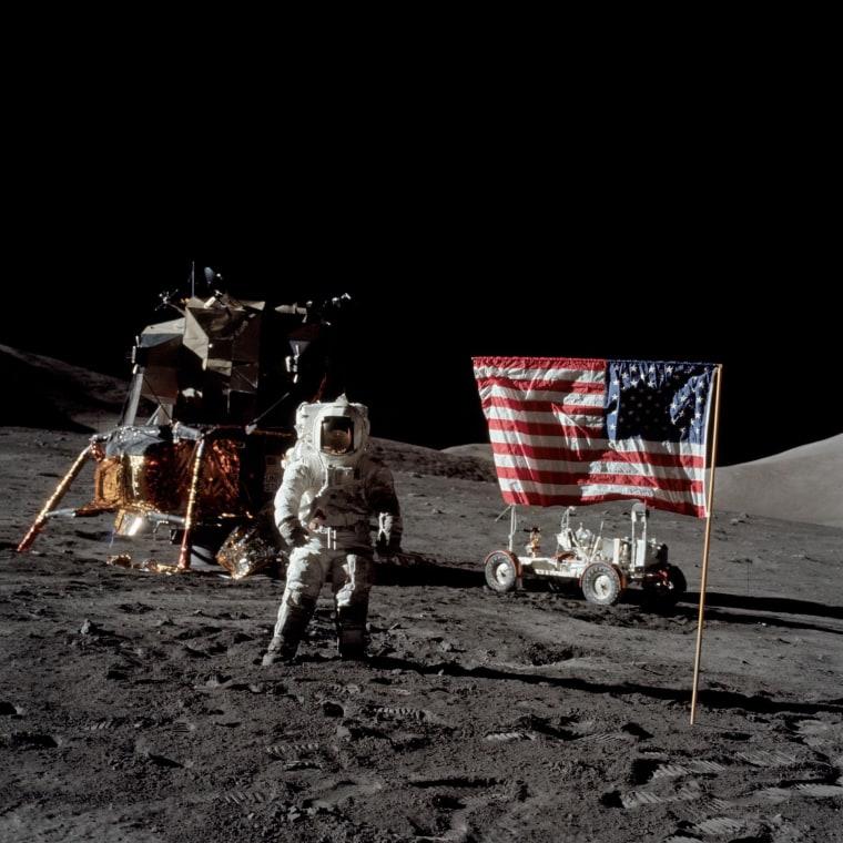 Harrison Schmitt stands on the moon in a space suit, with an American flag draped over his right hand, and a lunar module pilot on his left.