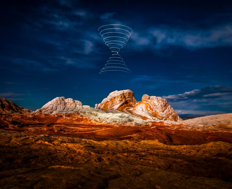 A rocky, desert landscape under a blue, cloudy night sky. In the sky above the rock formations, a spiral pattern of white lights forms the shape of two cones, similar to a vortex or an hourglass.