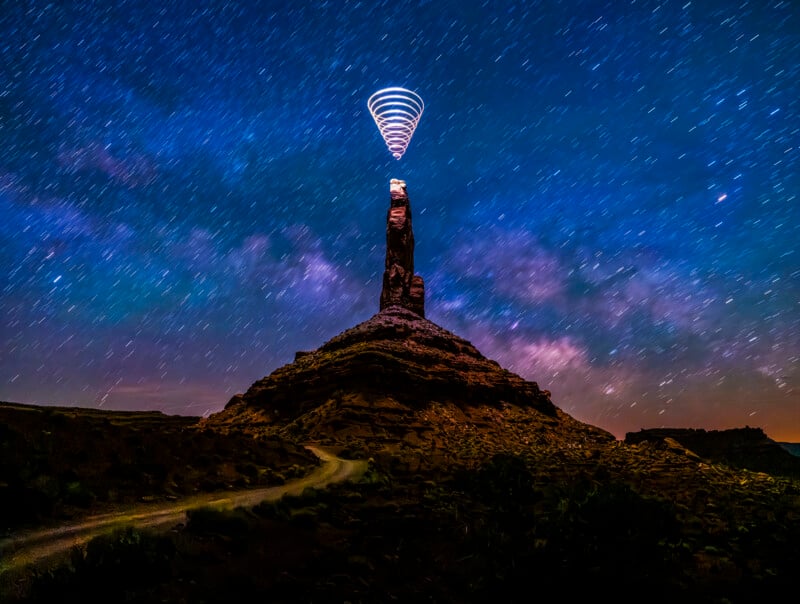 A tall rock formation stands under a starry night sky, with long luminous trails forming a cone shape on top of it, creating a surreal, luminous effect. A winding road leads to form.