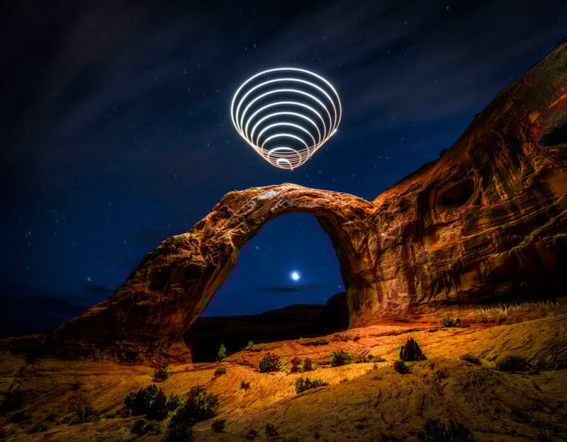 A natural tablet under a starry night sky, with spiral light trails above it, to create a futuristic effect. The landscape is illuminated, highlighting red rock formations and scattered vegetation.