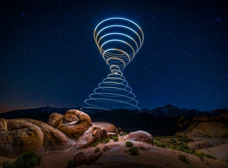Rocks and bushes under the night sky and distant mountains; spiral patterns of light trail up into the air, creating a bright vortex effect on the surface of the land.