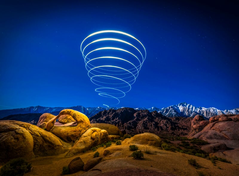 Rocky desert landscape under a blue sky at night, with mountains in the background. Bright spiral light trails rise above the rocks, forming a mysterious pattern above.