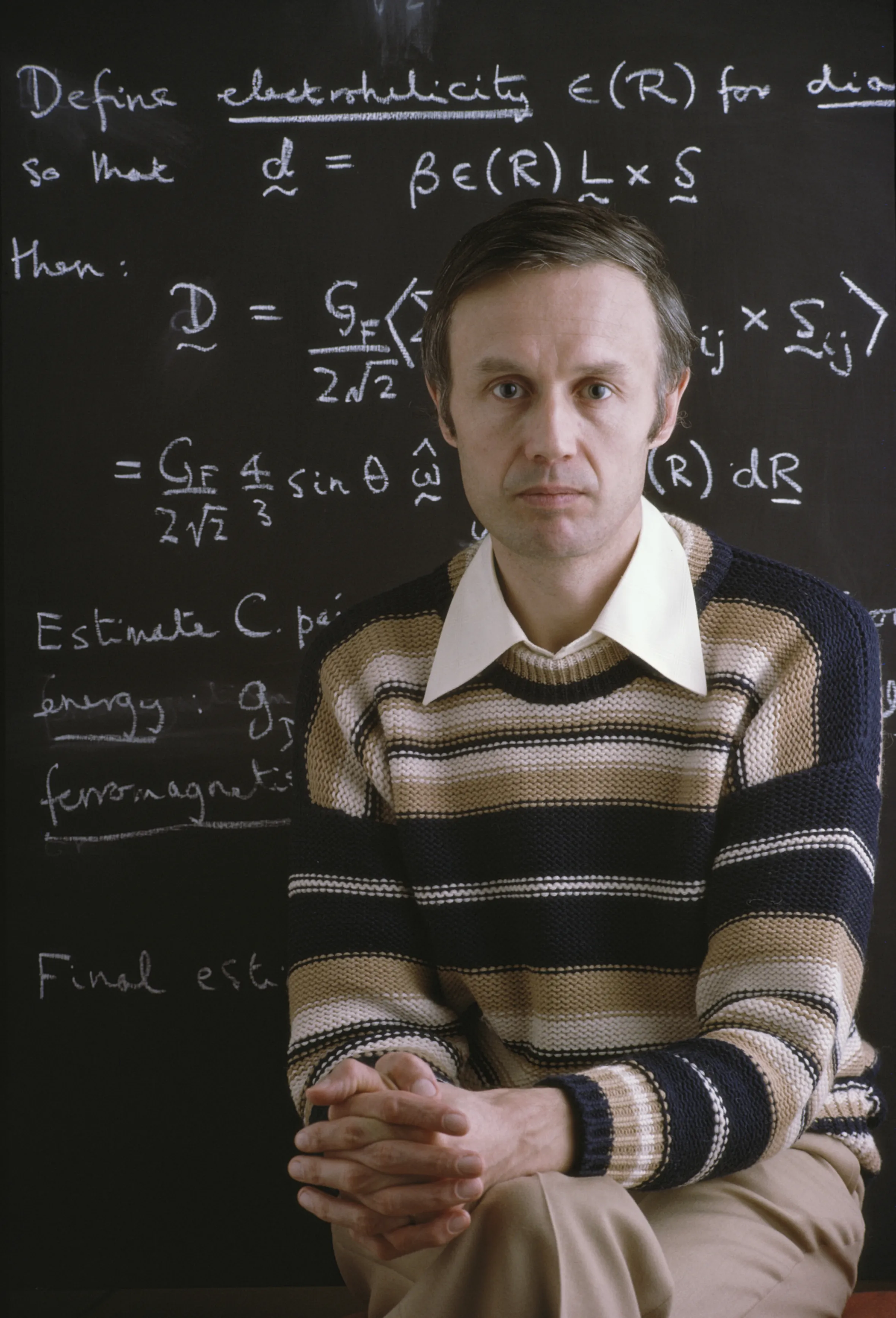 Sir Anthony James Leggett, physics professor and Nobel Prize laureate, sits in front of a blackboard covered in physics calculations.