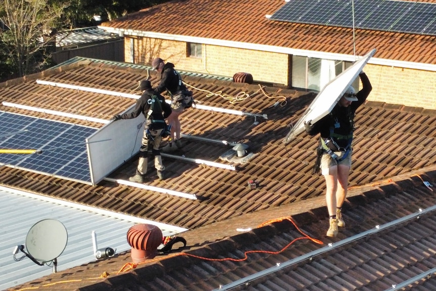 Men in dark clothes stand on a terracotta-tiled roof pulling out solar panels.