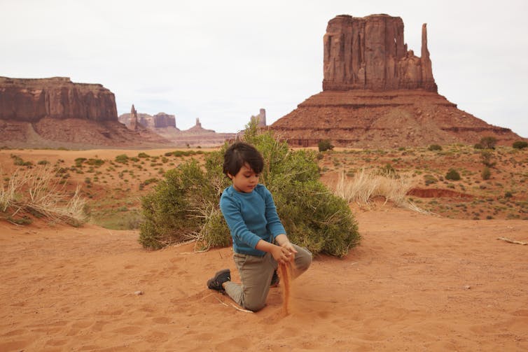 A child plays in the sand in front of a rock in Monument Valley