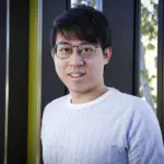 A smiling man wearing round glasses and a light blue sweater, photographed in front of a dark frame window.