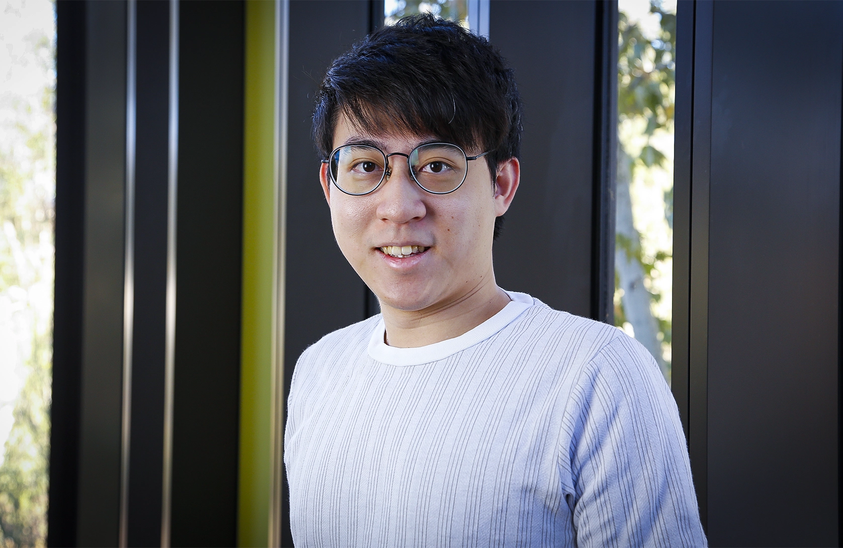 A smiling man wearing round glasses and a light blue sweater, photographed in front of a dark frame window.